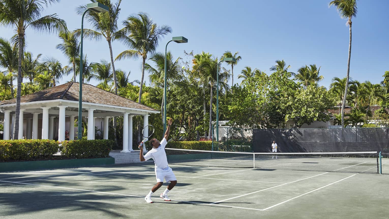 A man in all white trows a tennis ball high into the air, mid-serve 