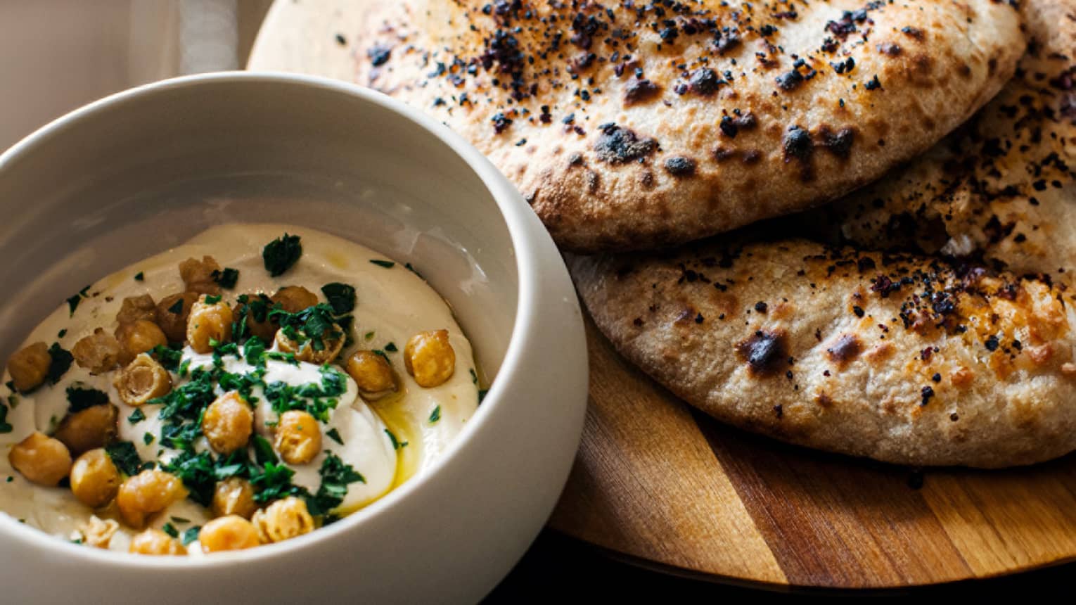 A white bowl of cream-coloured dip with garnishes and a wood cutting board holding pieces of round, flat bread
