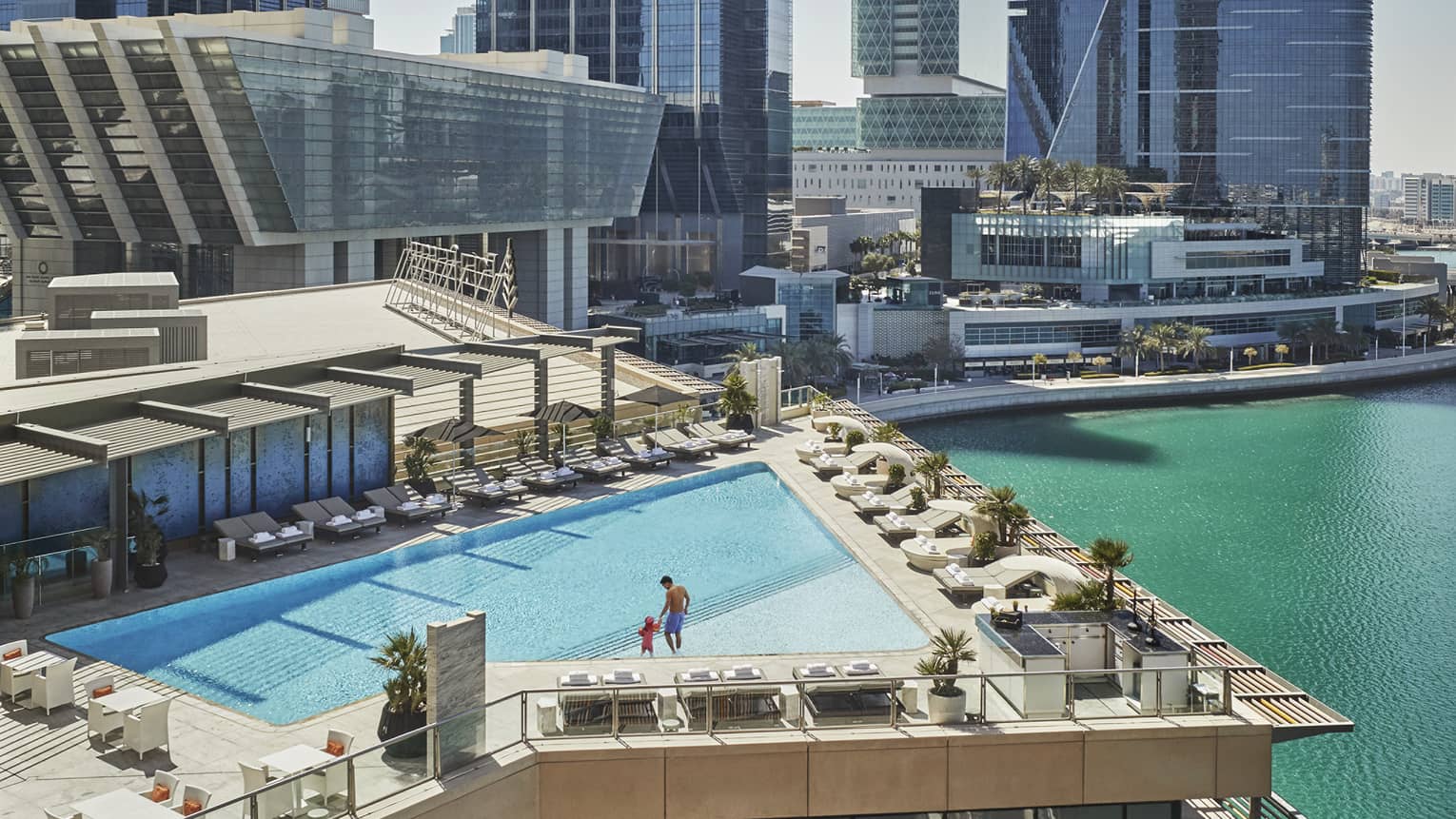 A wide shot of a man and child walking around a triangular pool overlooking the water and Abu Dhabi skyscrapers in the background. 