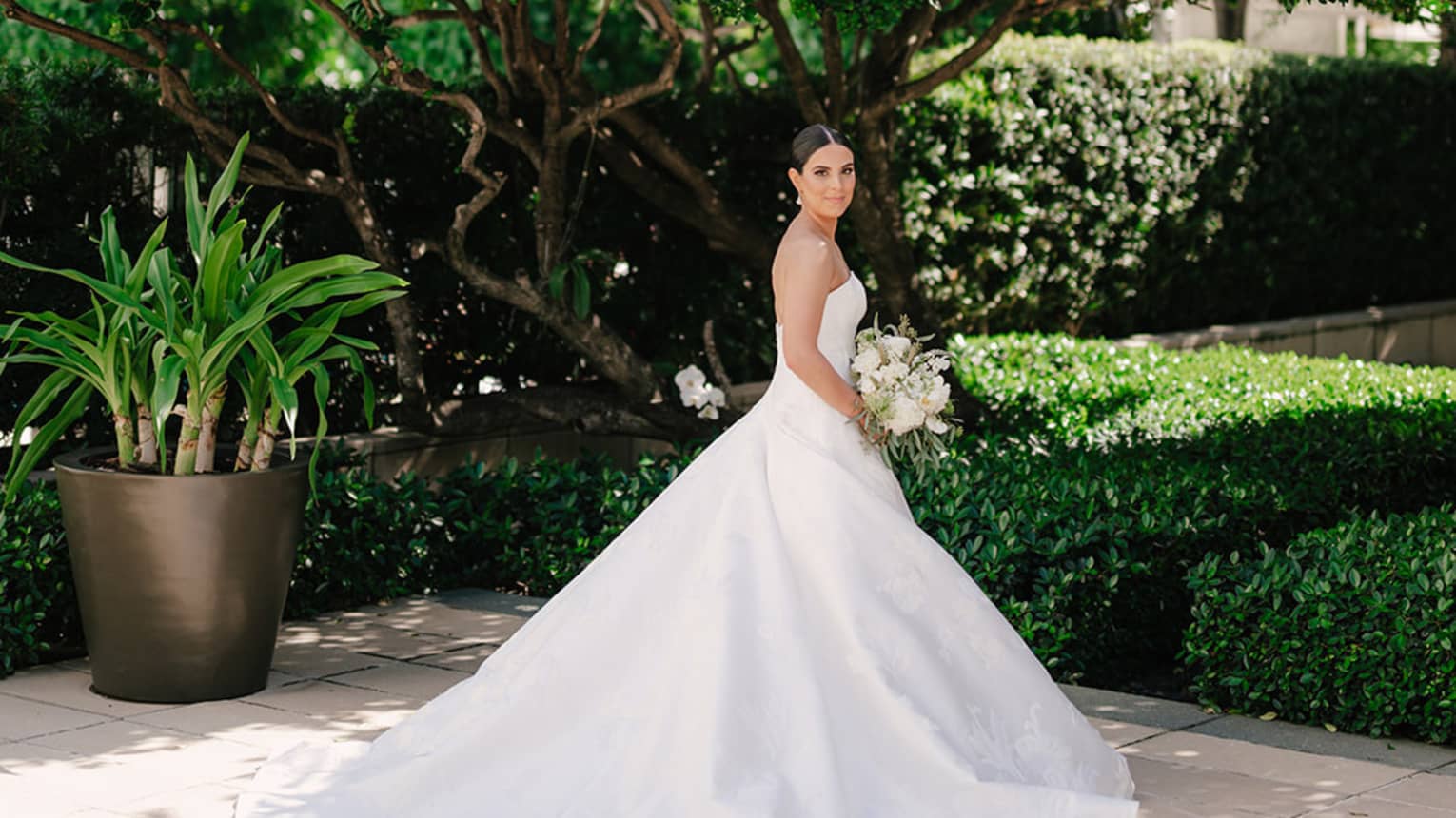 Bride in white ballgown poses with a white floral bouquet beneath a tree in an outdoor garden