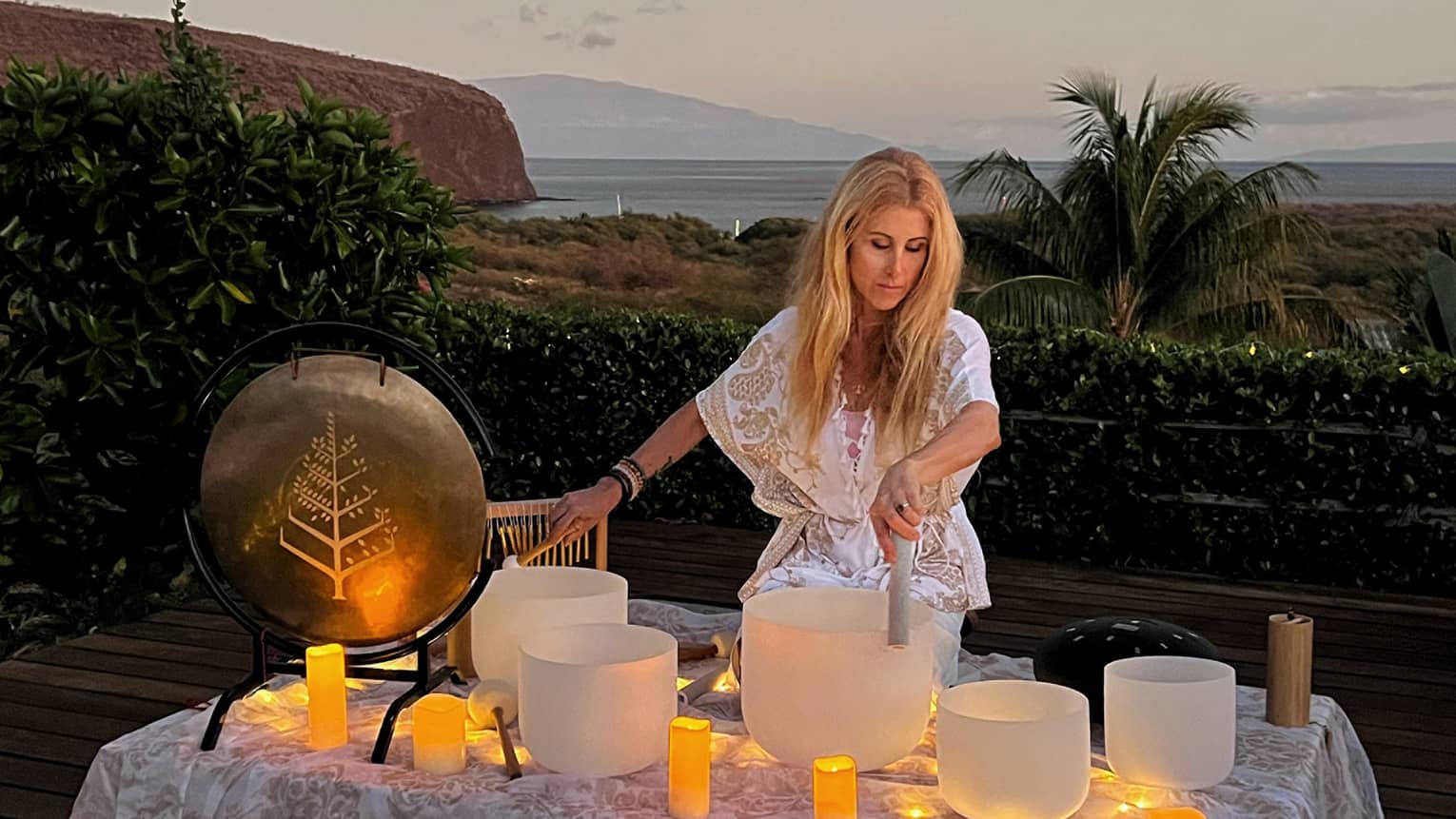 A woman performs crystal singing bowl ritual outside in beach setting at dusk