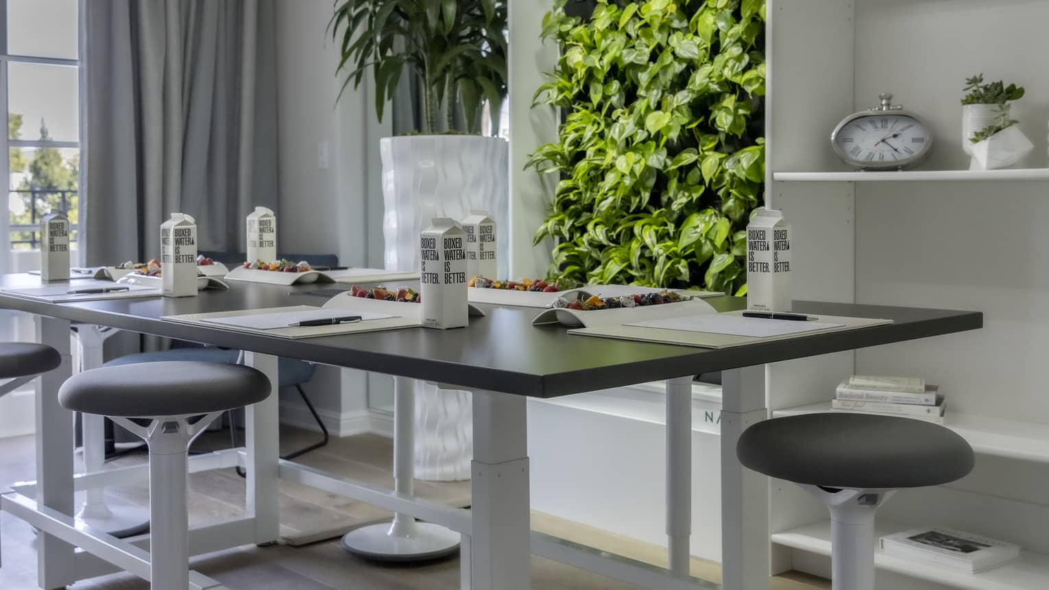 Stools surrounding a large desk with drinks and papers on it; with plants and shelves behind the desk.