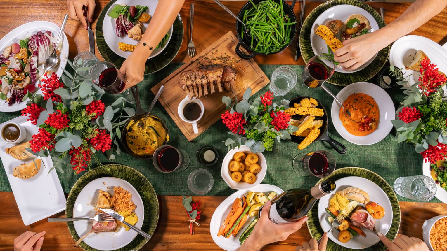 Wooden rectangular dining table topped with dark-green runner, three vases of greenery and red berries and four plates of food as arms reach around to table to serve their plates