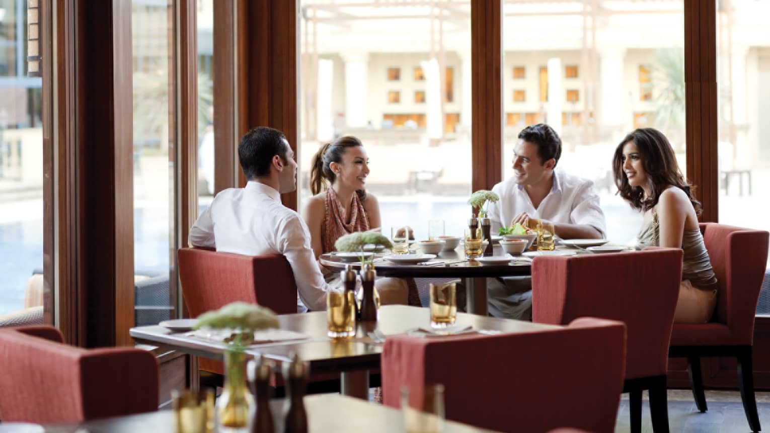 Four friends laugh, eat around Aura restaurant dining table by sunny corner windows 