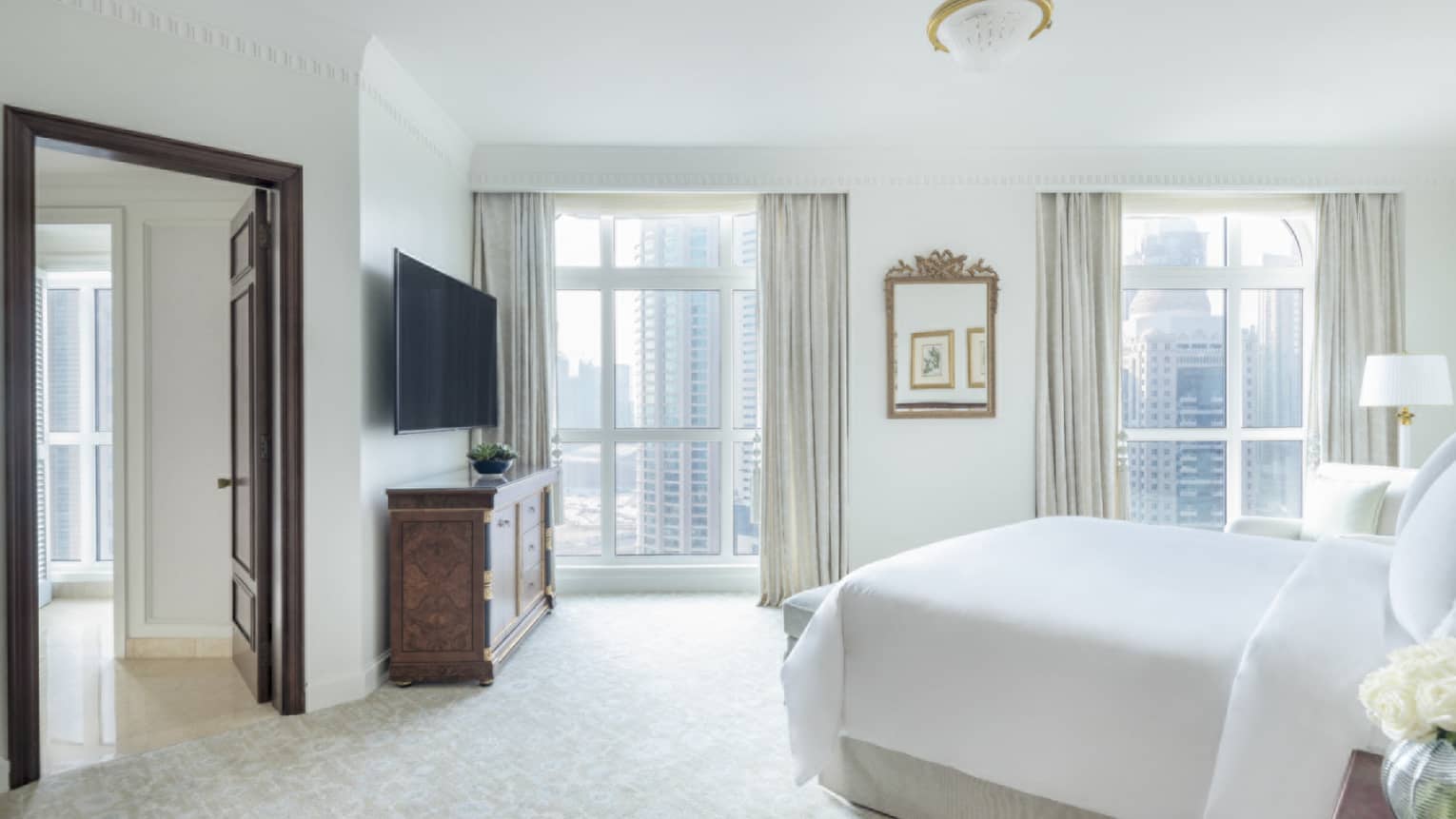 Light-filled bedroom with white bed, two floor-to-ceiling windows looking out to Doha city