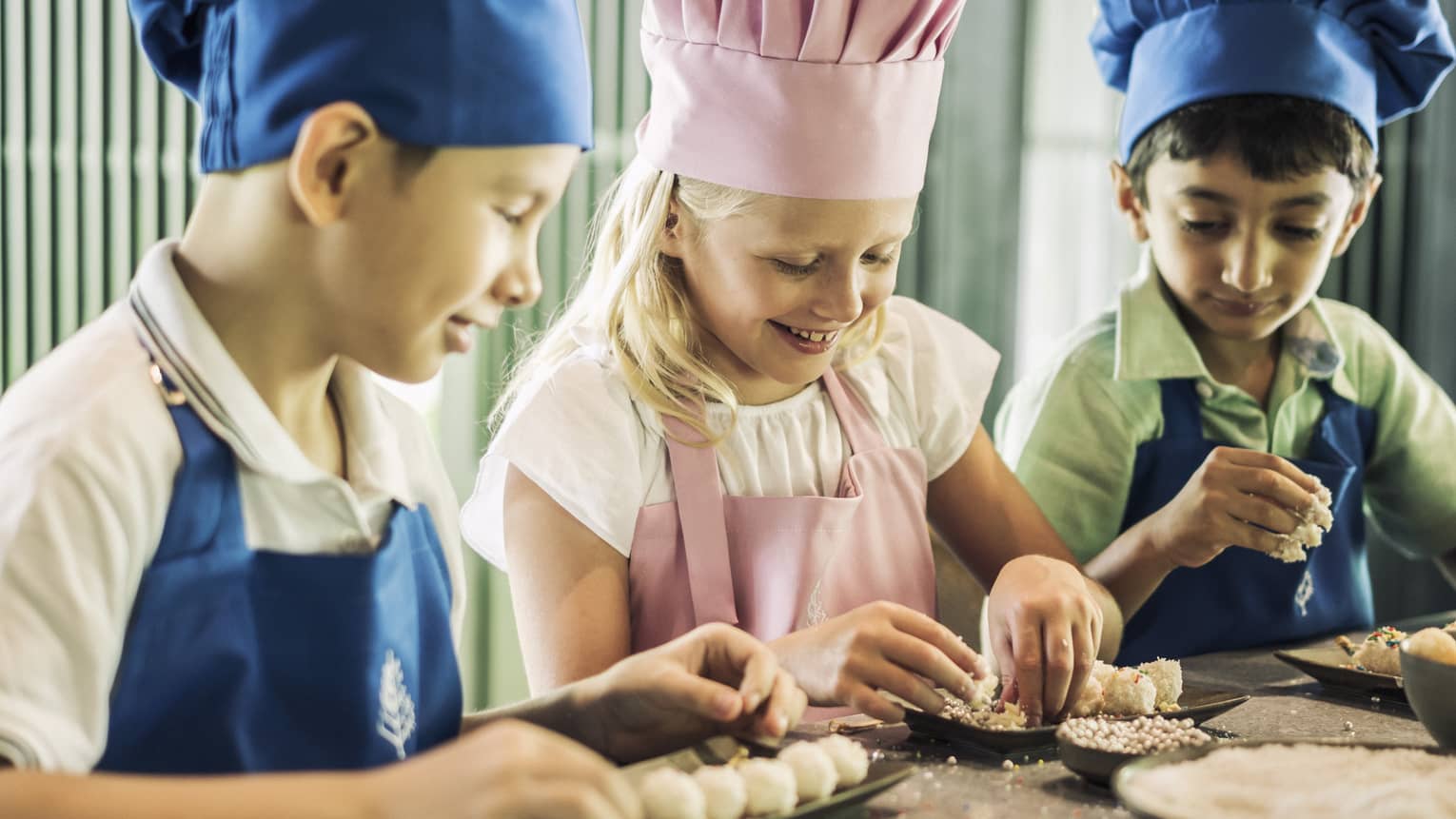 Three children in blue and pink aprons and chef hats roll pastries on small black trays