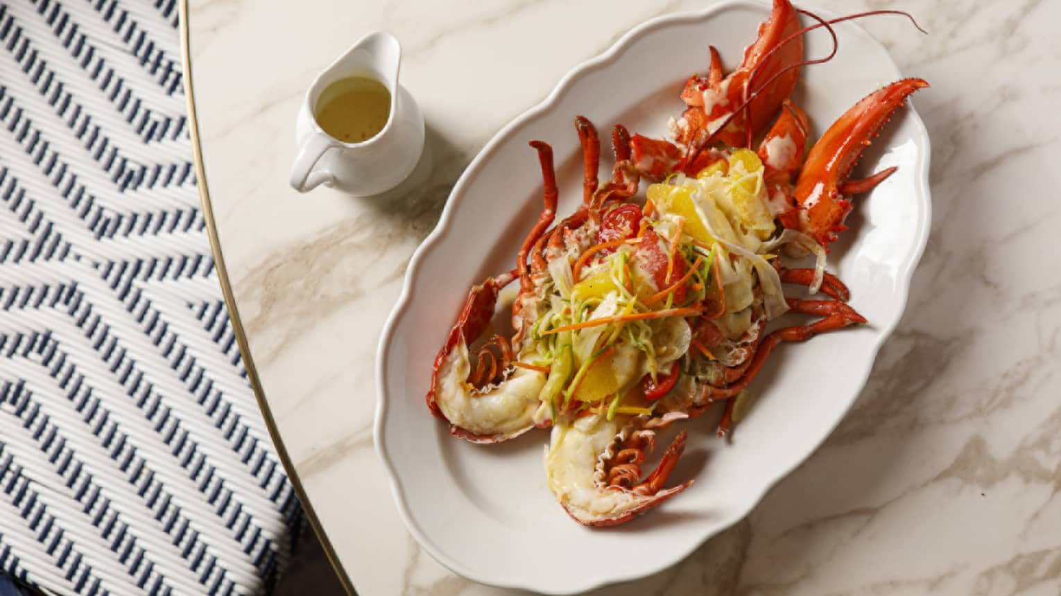 An oblong white platter with a scalloped edge holds a lobster preparation. A small pitcher of sauce is next to the platter.