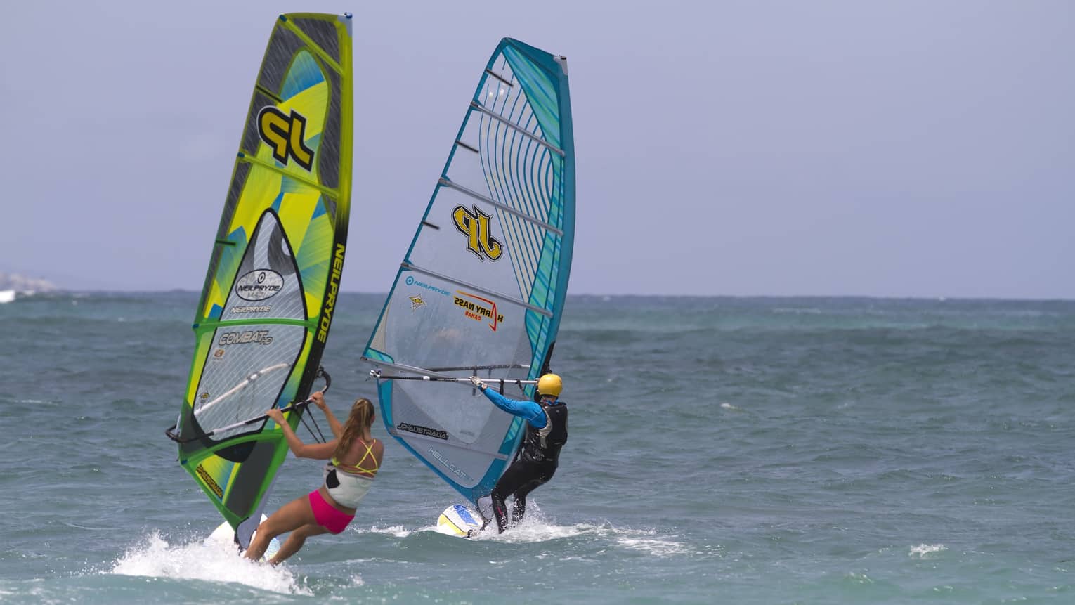 Two windsurfers leaning into the open sea under an azure blue sky, each creating a small wake.