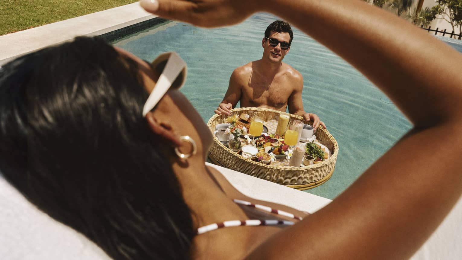 A guest stands in a pool with a floating basket of breakfast food and drinks and looks up at another guest sitting poolside.
