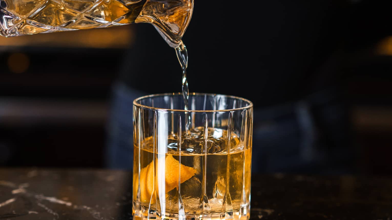 An amber cocktail being poured from a decorative clear vessel into a clear tumbler
