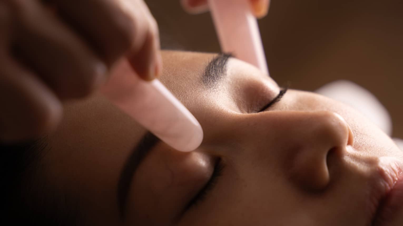 Hands hold pair of pink jade stones against woman’s eyelids during facial