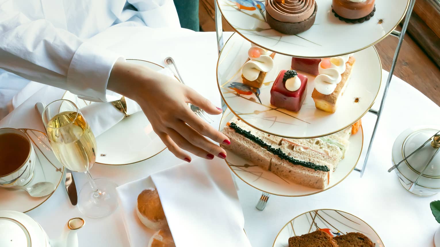 Person in white shirt reaches to classic afternoon tea stand filled with patisserie and mini sandwiches