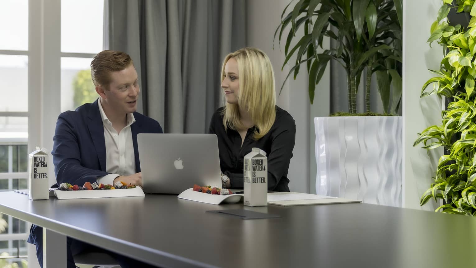 A man and woman in business clothes using a laptop and discussing something while at a table.