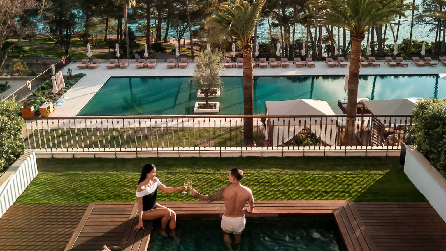 Two people in a small square pool in an outdoor setting. A larger pool surrounded by lounge chairs is in the background, as are ocean and mountain views beyond that. Verdant lawns and trees surround the pool areas.
