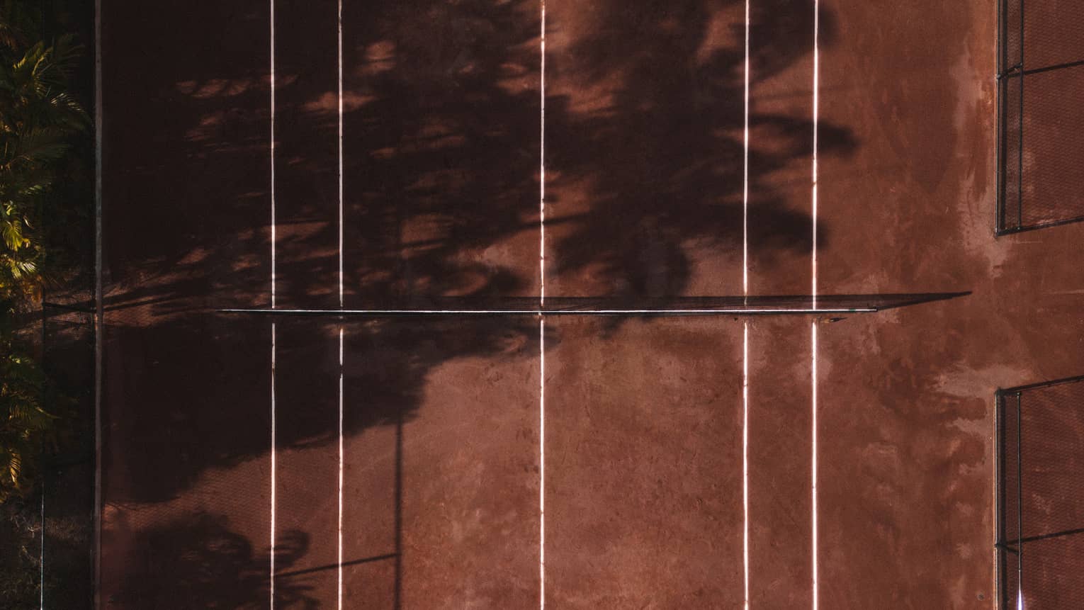 An aerial view of a red clay tennis court partially shaded by trees