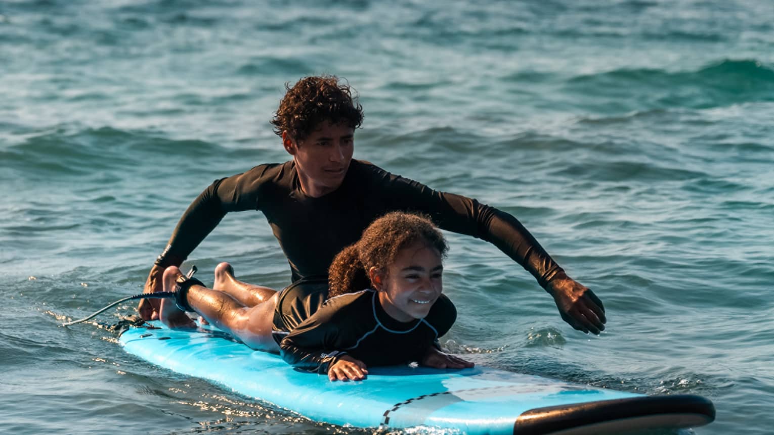 Male suf instructor wearing a black long-sleeve rashguard helps a young surfer paddle in the ocean