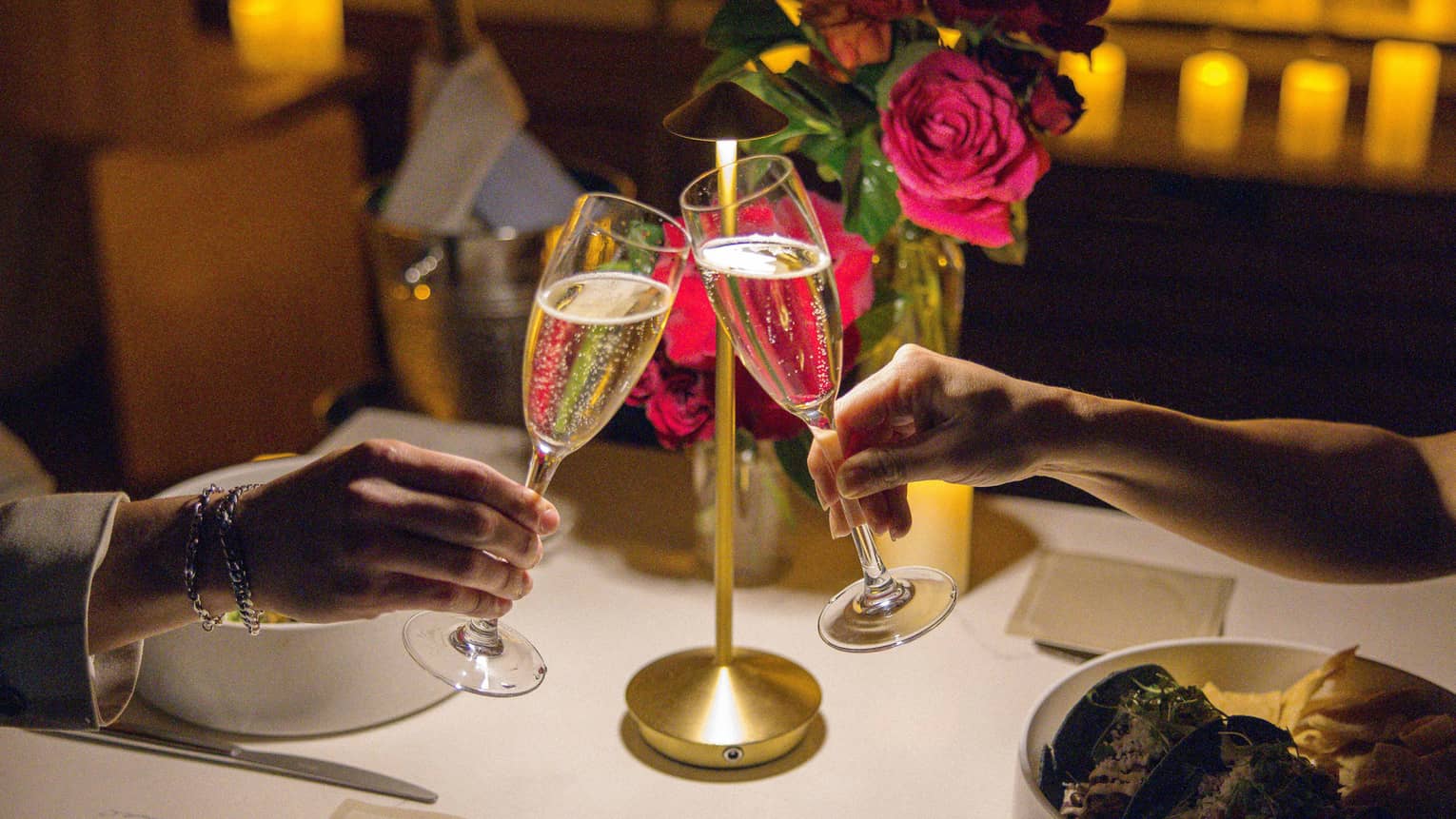 Two hands clink champagne glasses over a dining table set with flowers, candles in the background