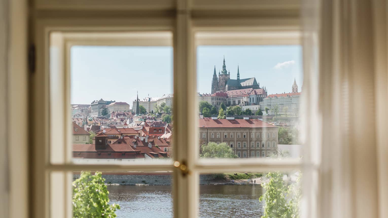 View through a window of a historic city with red rooftops and a cathedral in the distance, overlooking a river and green trees under a clear blue sky.