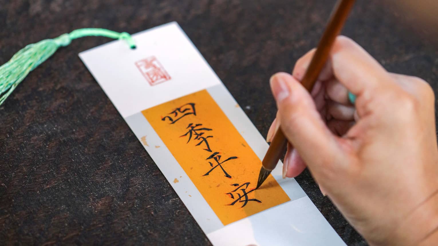 Chinese calligraphy being written in black ink onto a gold banner on a crisp white paper bookmark with a mint-green tassel.