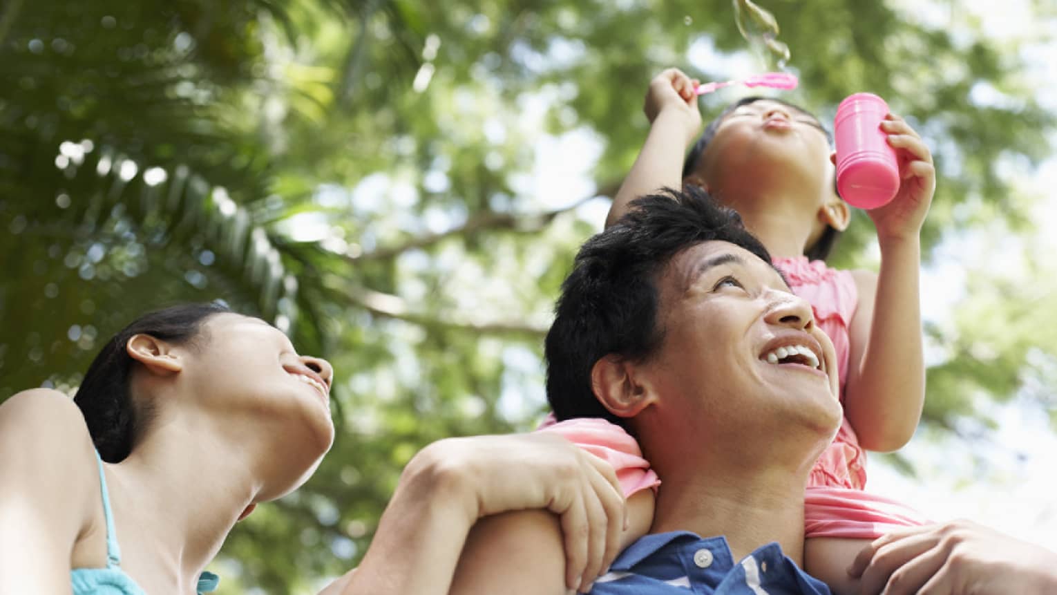 Young girl blows bubbles with pink wand as she sits on laughing man's shoulders, smiling woman beside him