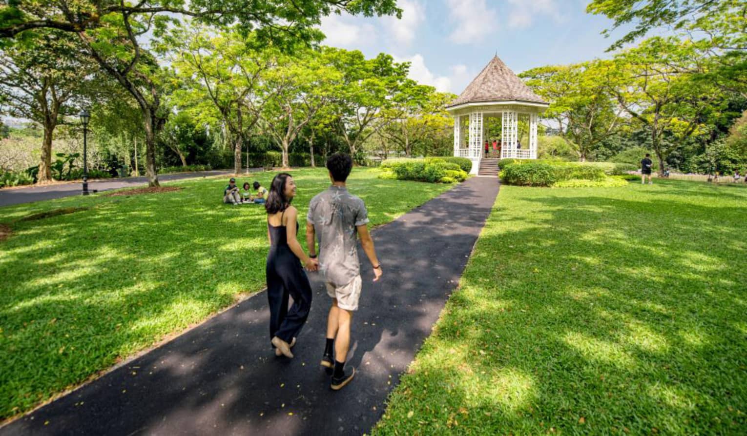 A man and woman hold hands and walk along an asphalt trail through the Singapore Botanic Gardens 