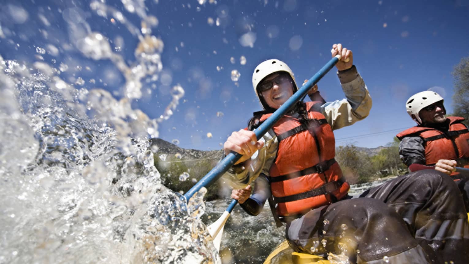 Smiling woman wearing red life vest holds paddles as waves crash over raft