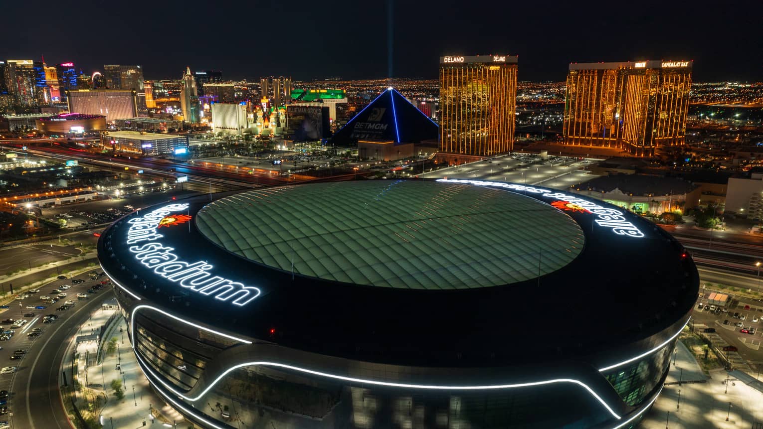 A nighttime, birds-eye view of a large sports stadium, with other tall buildings and lights around it.
