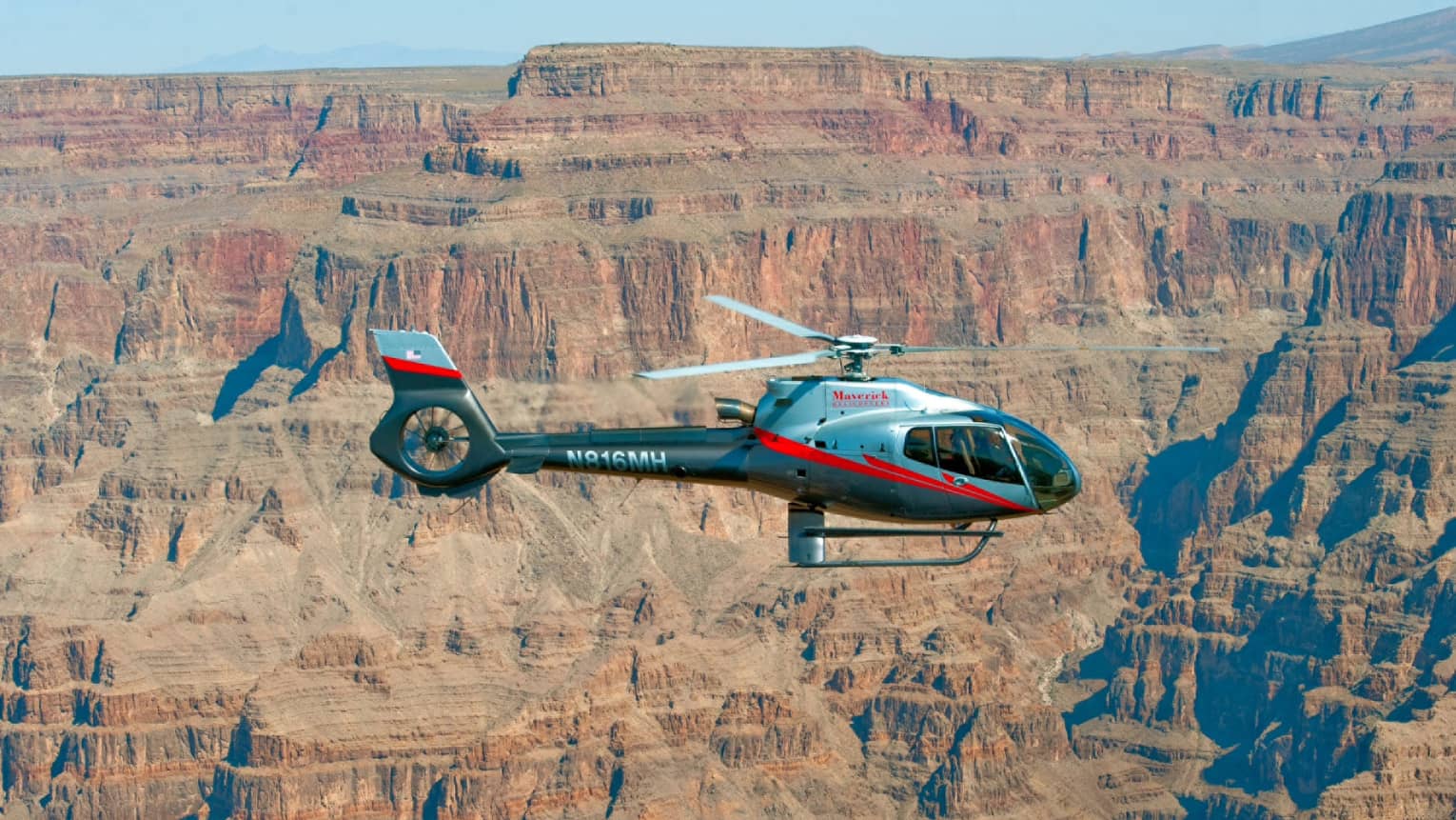 Maverick Helicopter Tours helicopter flying in sky in front of canyon cliffs