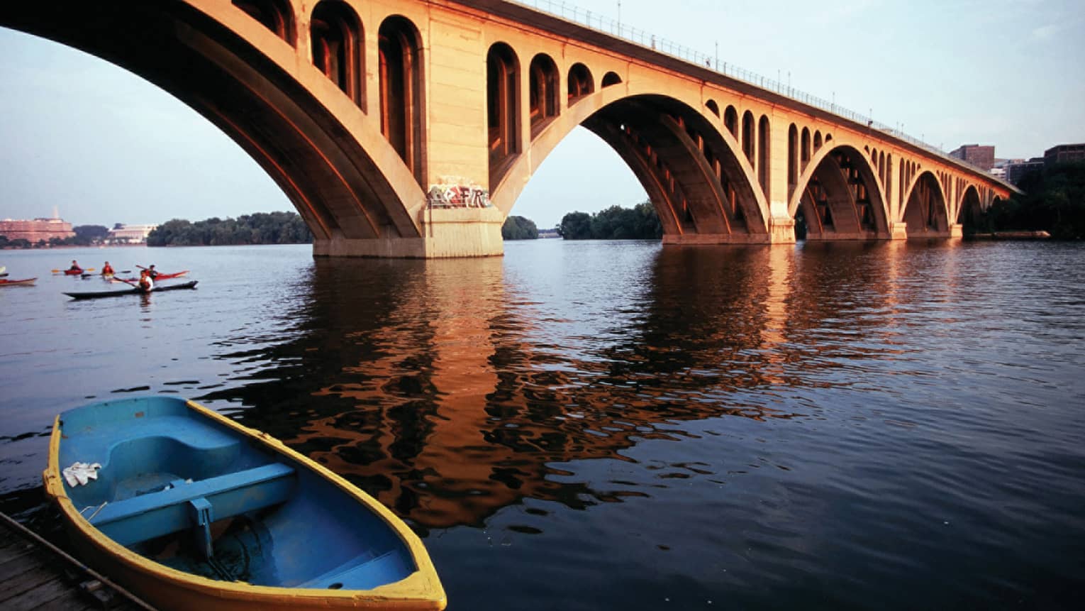 A wide river with a large bridge crossing it, there are people rowing kayaks under the bridge.