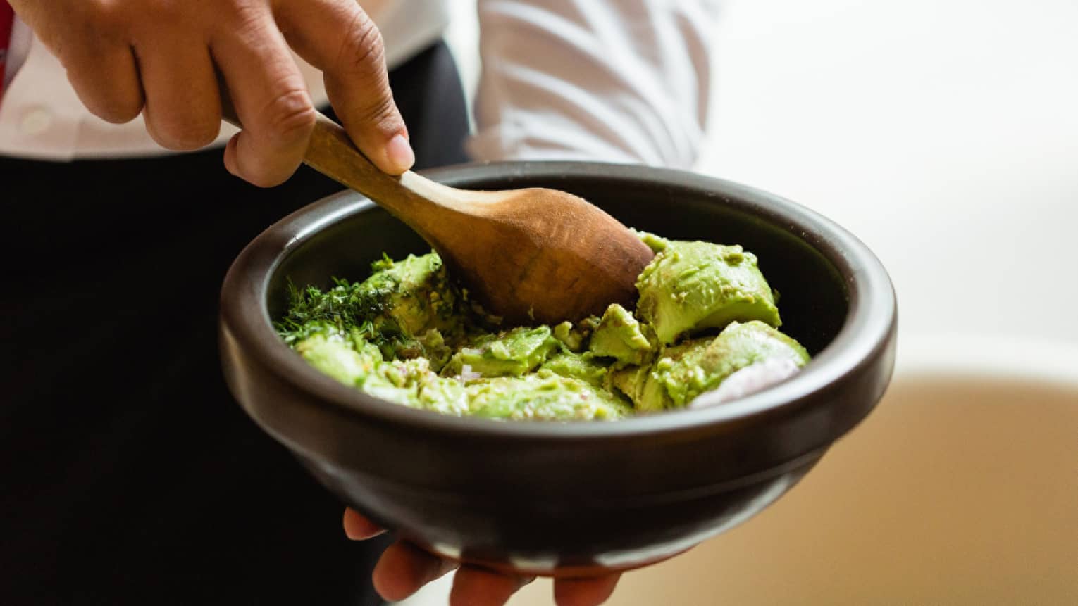 With a wooden spoon, a chef mashes up bright green avocado chunks with salt rocks and cilantro in a palm-sized black bowl. 