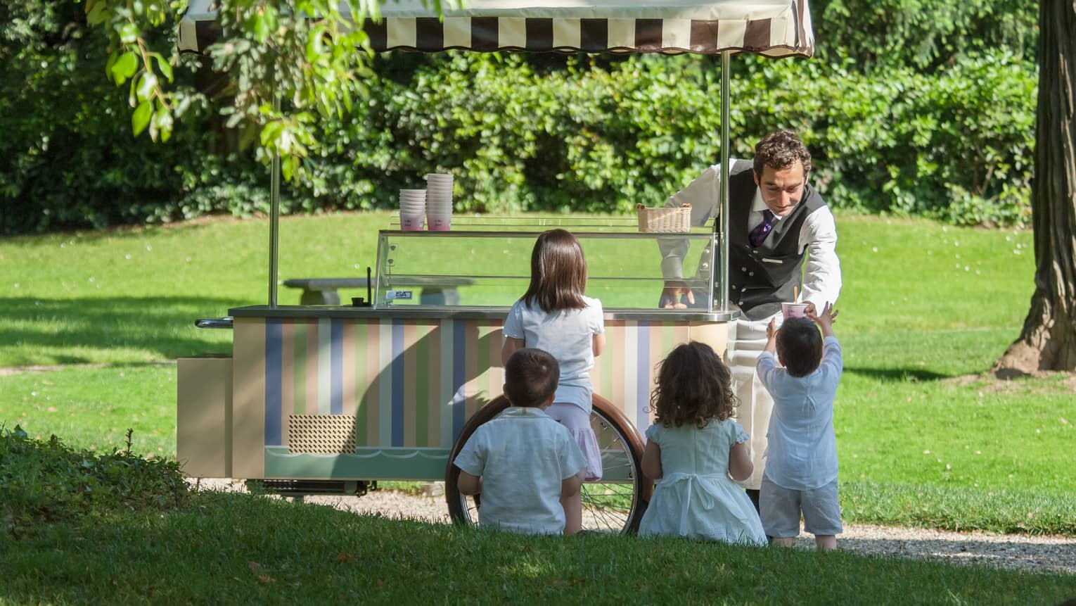 Man behind dessert cart serves ice cream to young children in sunny garden