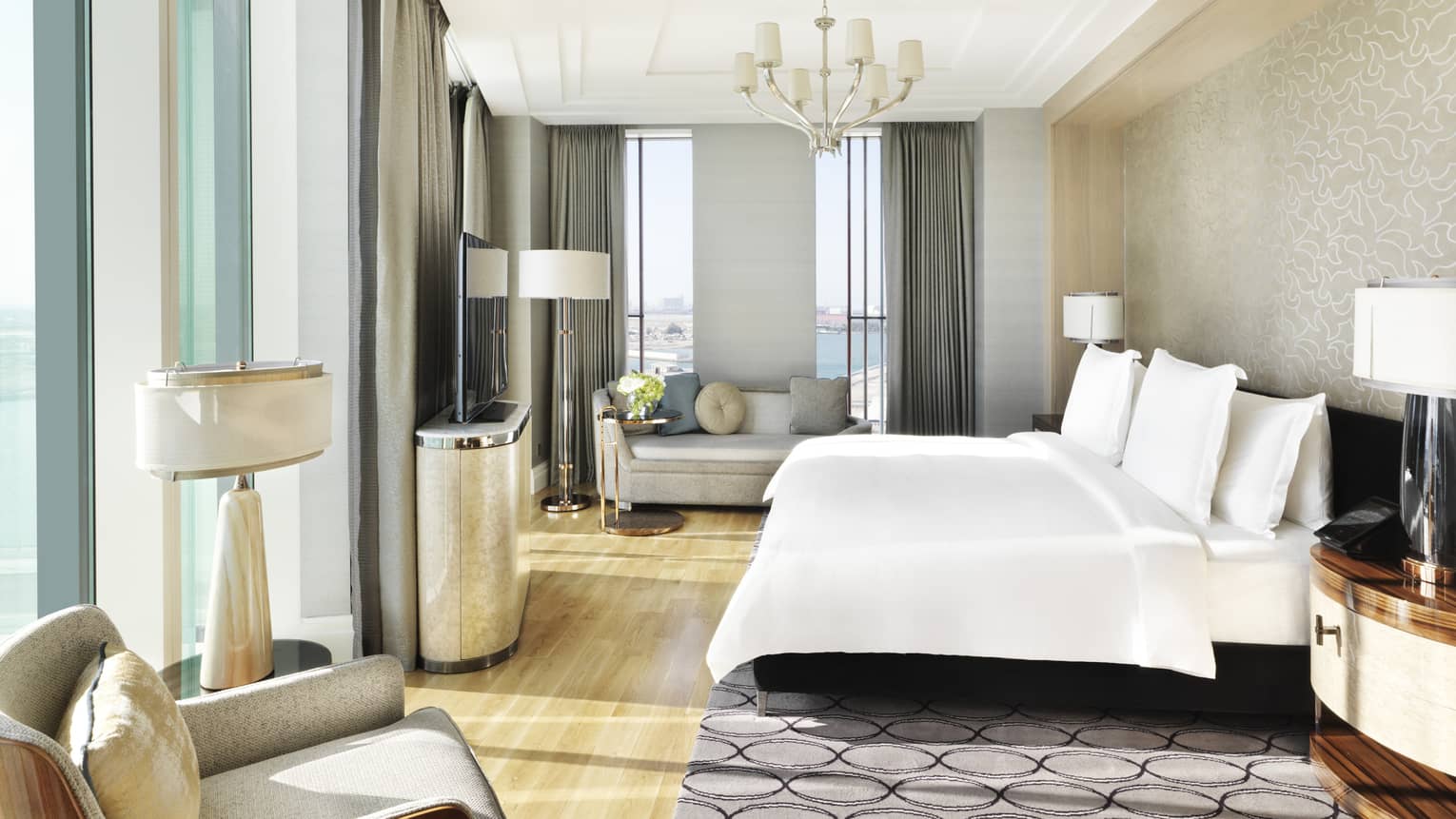 Executive Suite bed with white linens under chandelier, sunny corner windows, grey loveseat sofa and chair