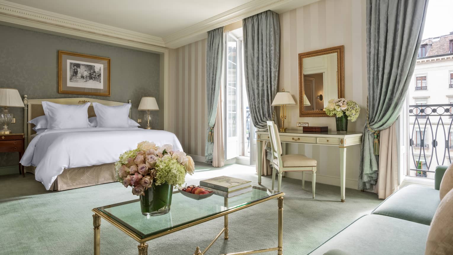 Junior Suite bed, glass coffee table with gold trim, white desk near French door and balcony, blue curtains