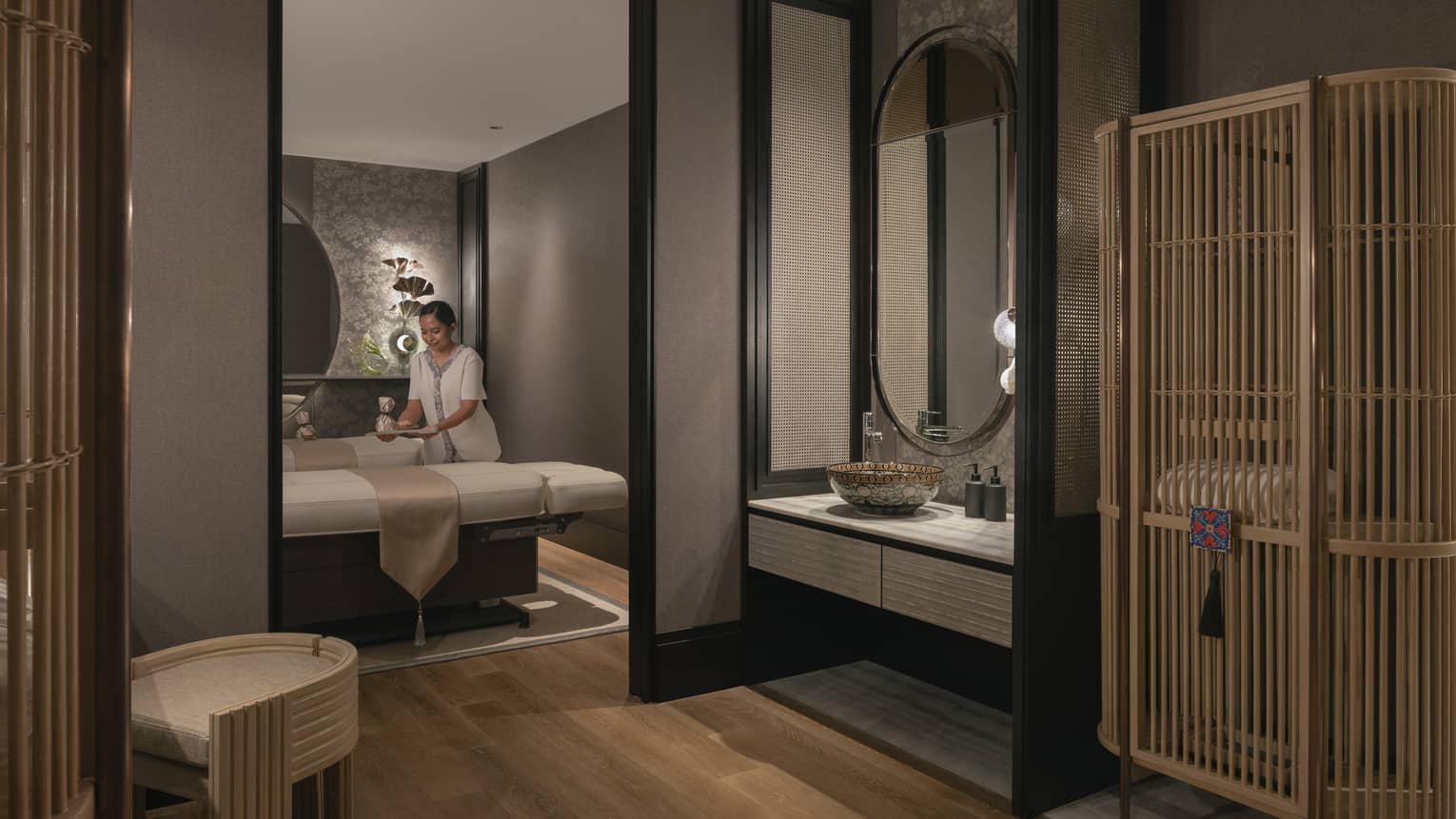 A tray is gently placed on one of two massage tables in a warmly lit spa room with soft grey walls and natural wood décor