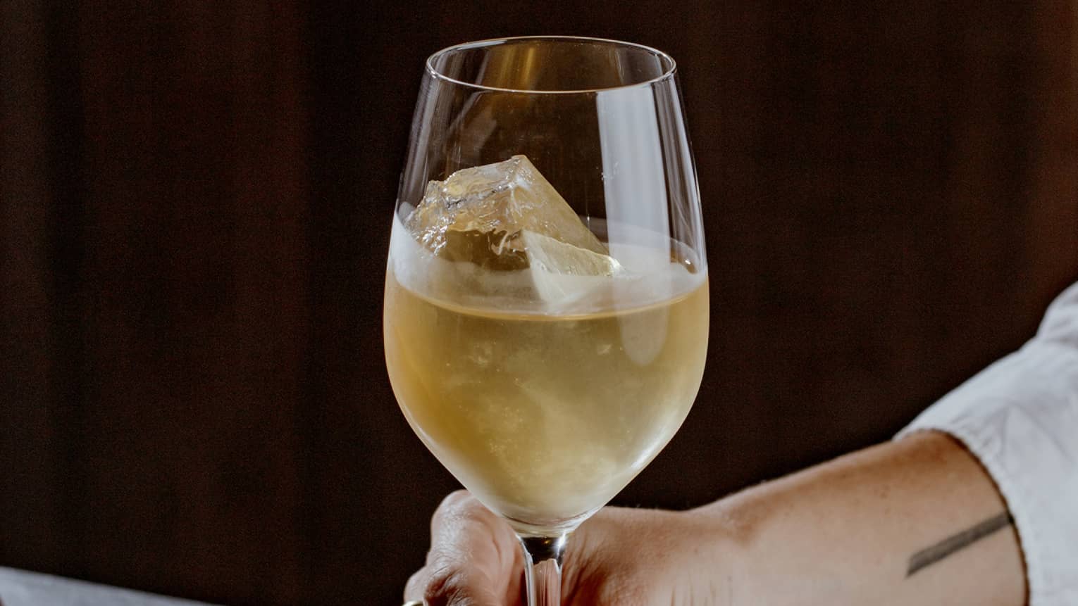 A hand holds onto the stem of a large cocktail glass filled with a pale golden beverage and ice