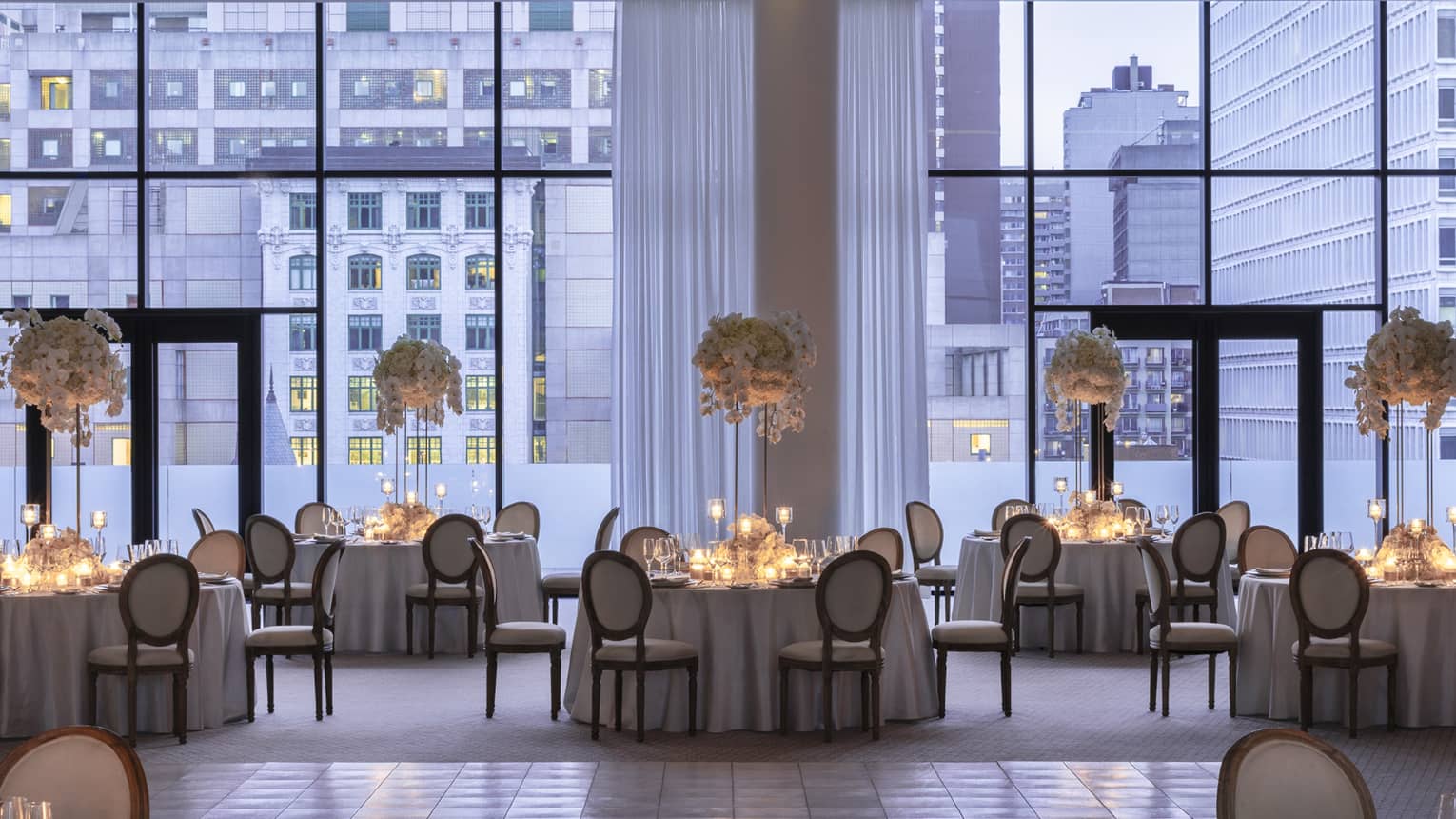Ballroom with floor-to-ceiling windows overlooking buildings, round tables with candles and large floral displays 