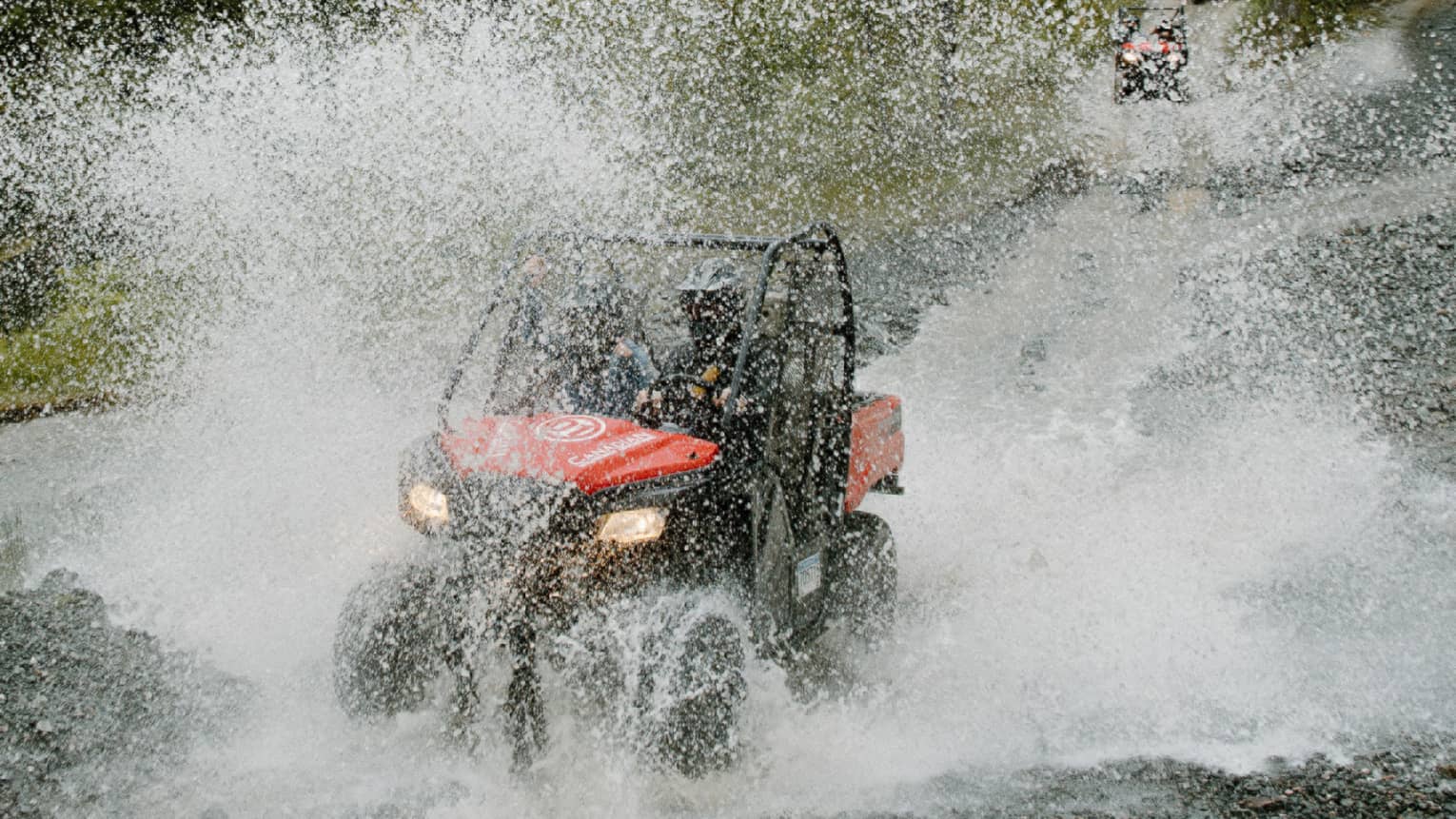 A red-and-black ATV driving through water, creating a thick veil of water spray against a background of pine forest.