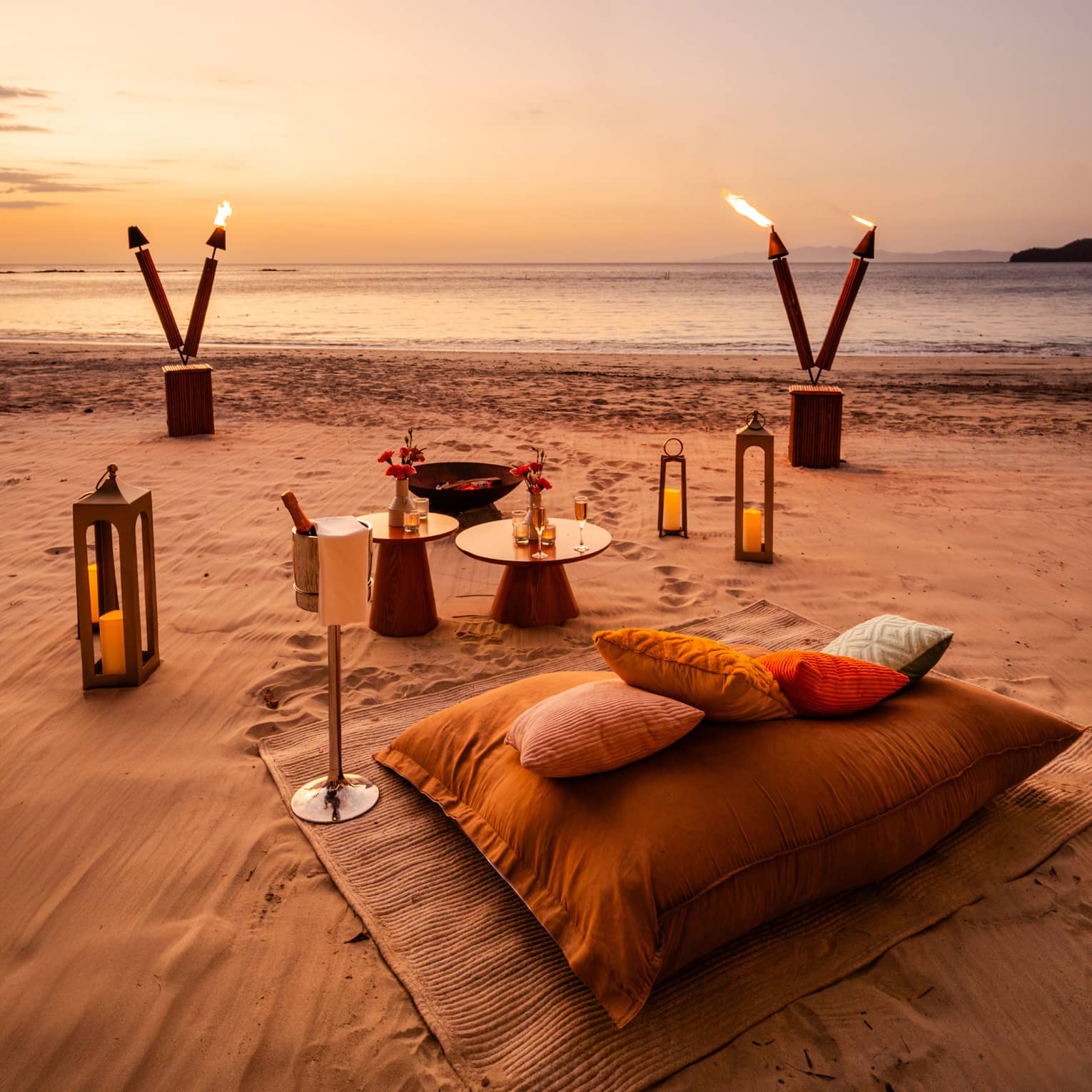 Blanket, cushions, small tables, lanterns and tiki torches are set up for a private dinner on a beach at sunset