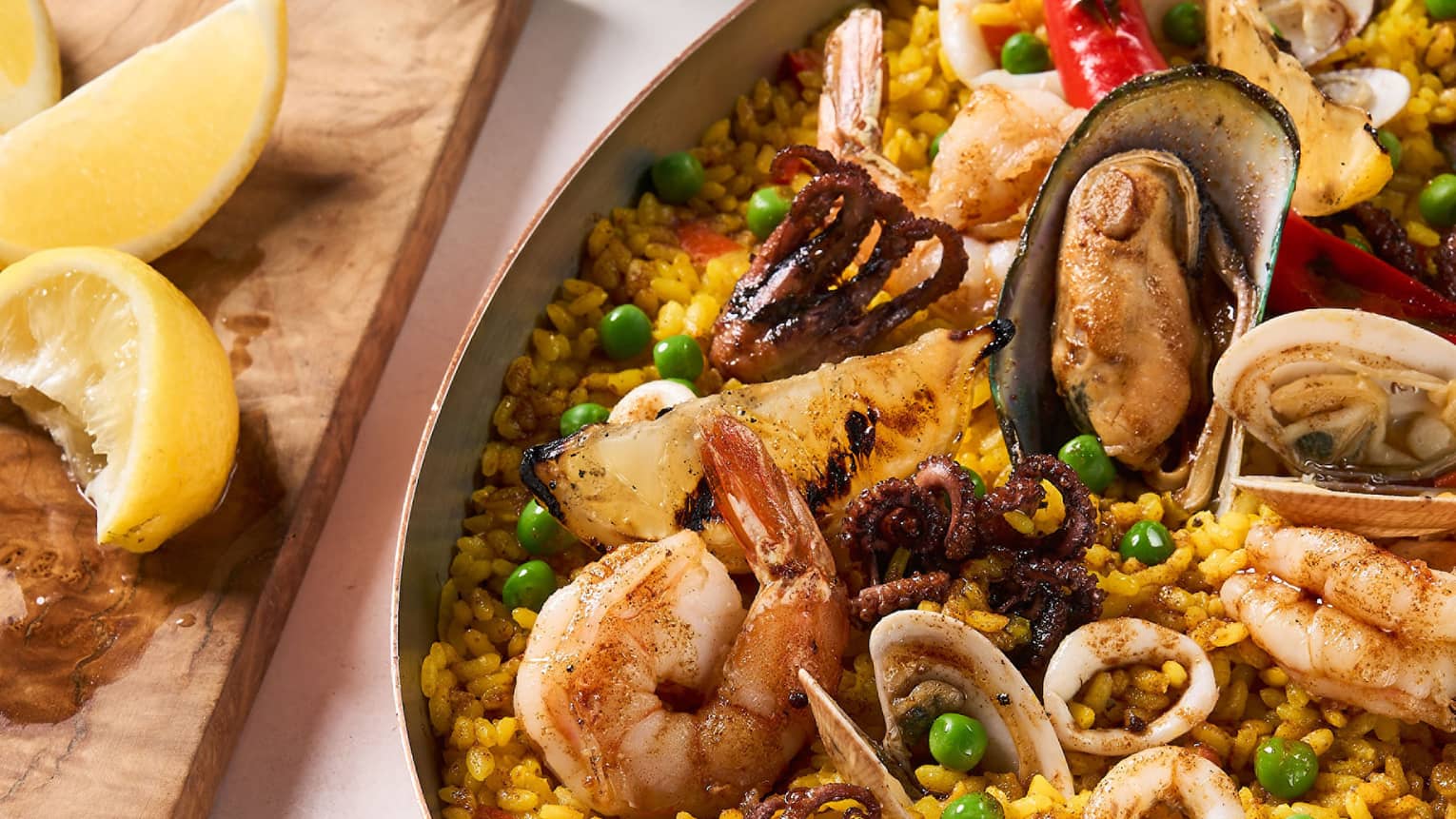 Seafood paella with assorted seafood in traditional oval-shaped metal pan, lemon wedges on cutting block