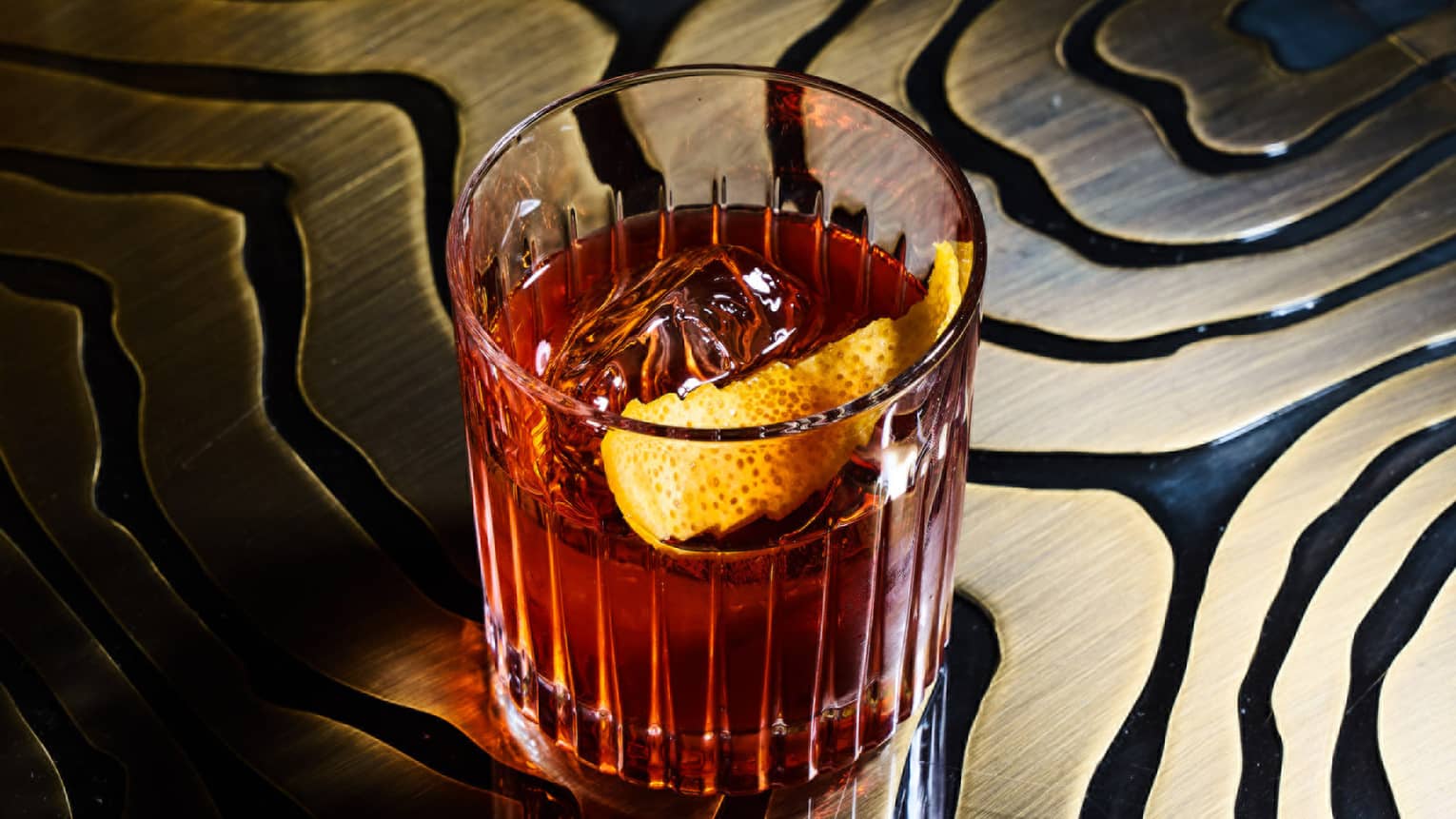 Old Fashioned cocktail with ice and orange rind in a clear Old Fashioned glass, sitting on a black and gold table