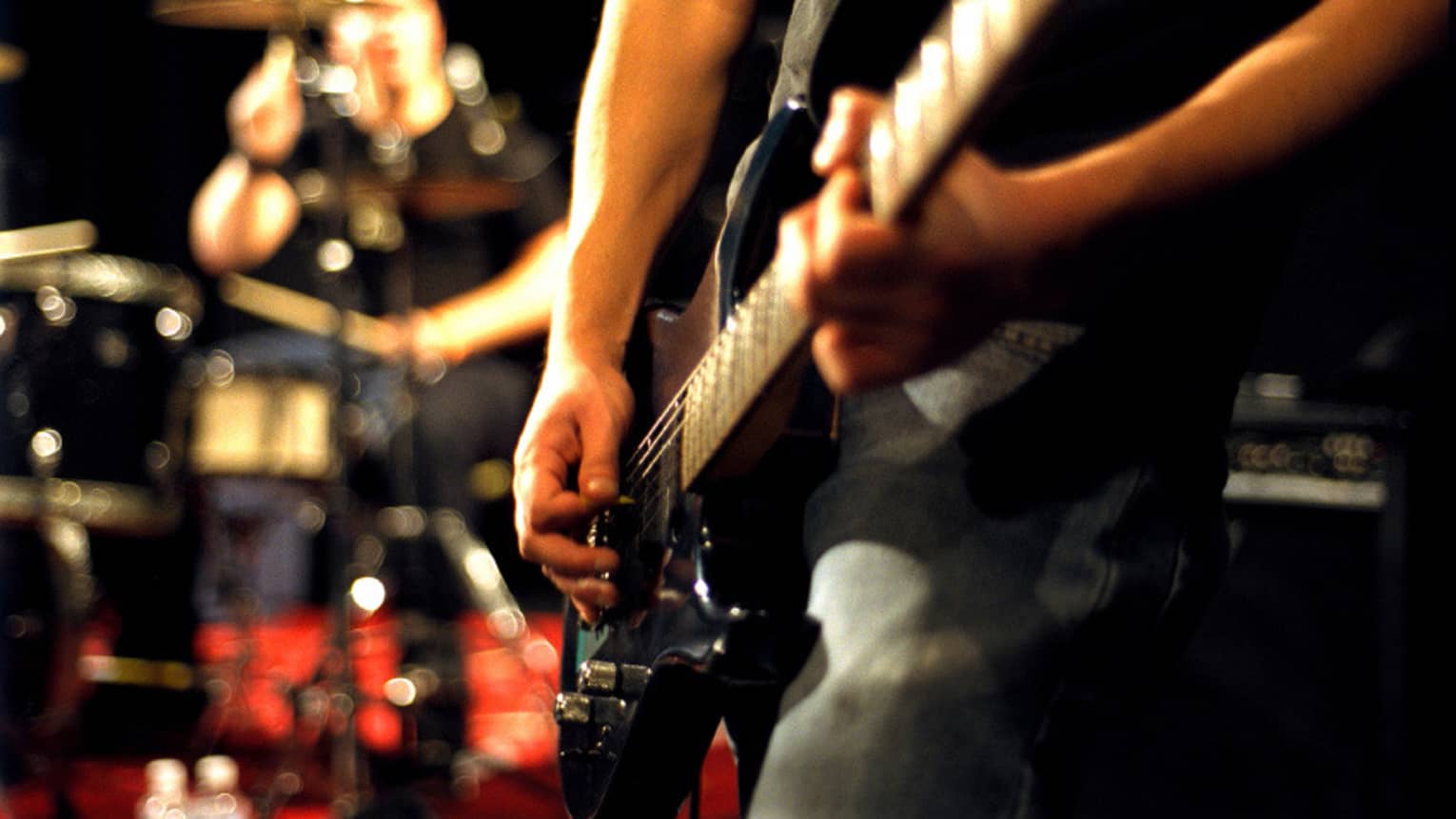 Close-up of musician's arms and hands playing an electric guitar with drumset in the background.