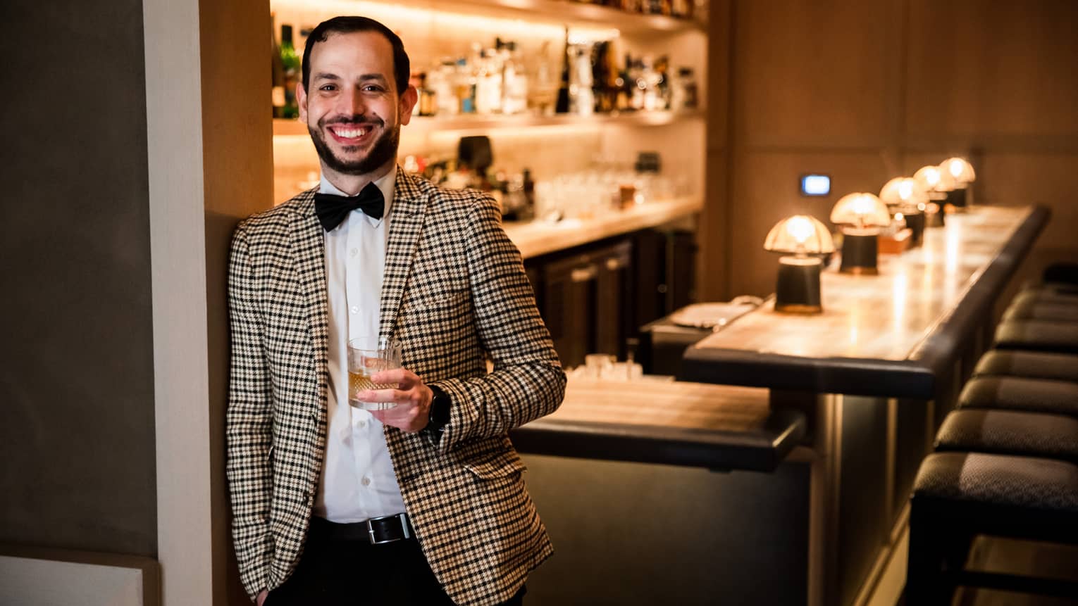A man in a checkered suit holding a cocktail in front of a bar.