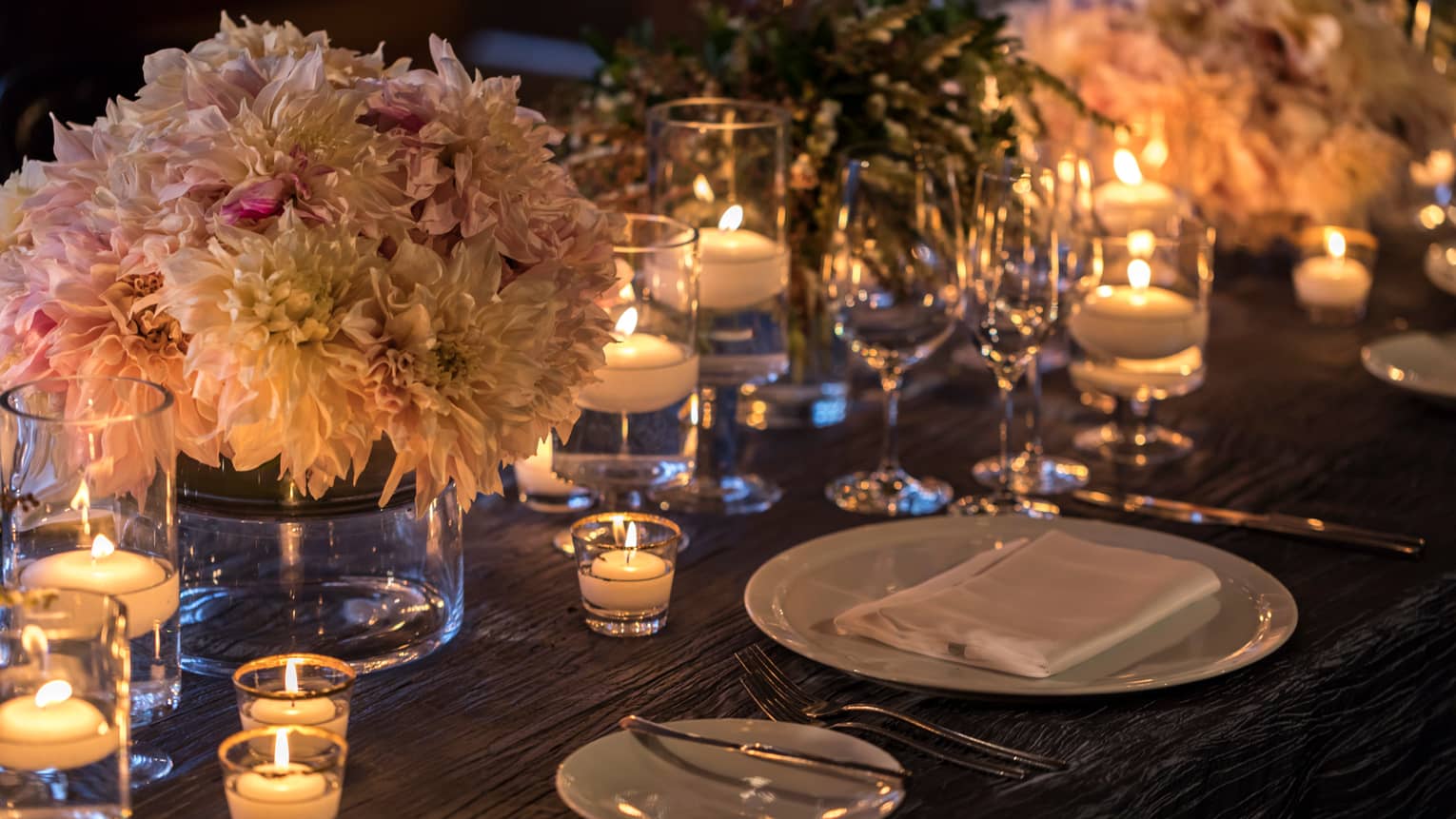 Pink and white flower arrangement in glass vase on dining table with glowing tealight candles