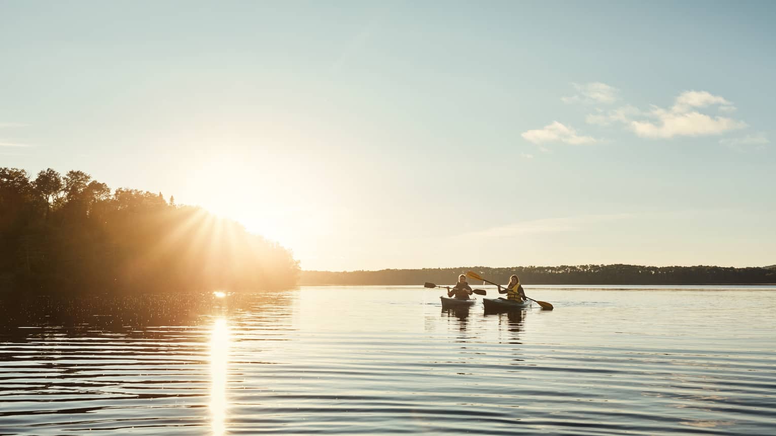 Two people kayaking through a body of water flanked by trees, both silhouetted by the setting sun