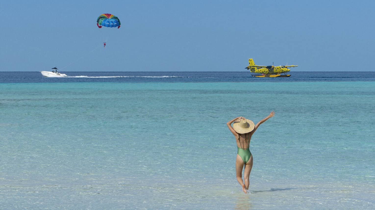 A woman walking on the beach watches a seaplane and a parasailor in the distance. 