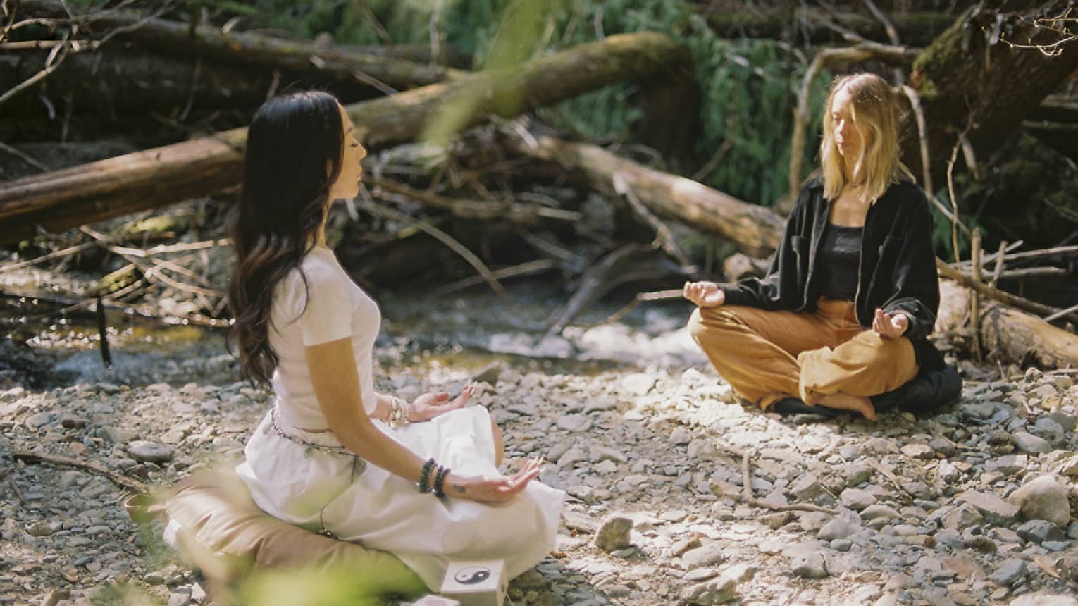 An instructor and a guest sit on yoga cushions as they peacefully meditate in a small clearing beside a brook in a forest.