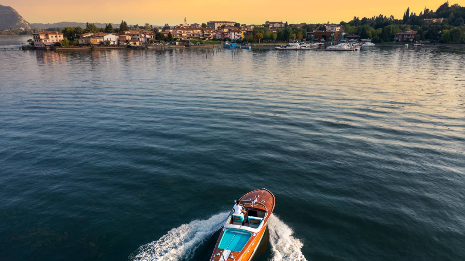 Person driving a wooden speedboat across a lake towards shore at sunset