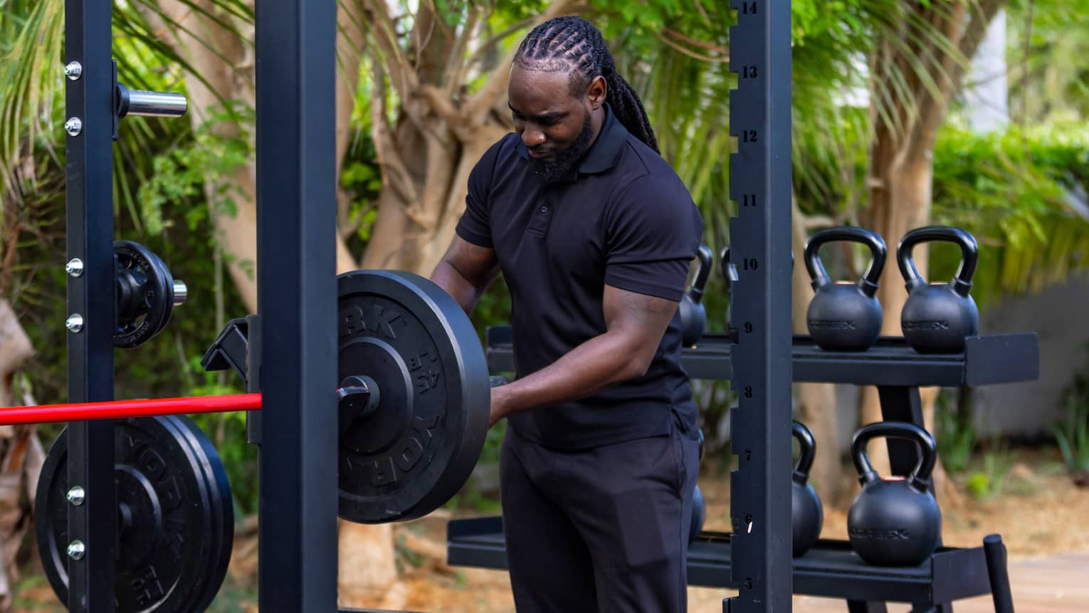 A guest adjusts a barbell in an outdoor gym against a rack of kettlebells and medicine balls and a grove of shady trees.