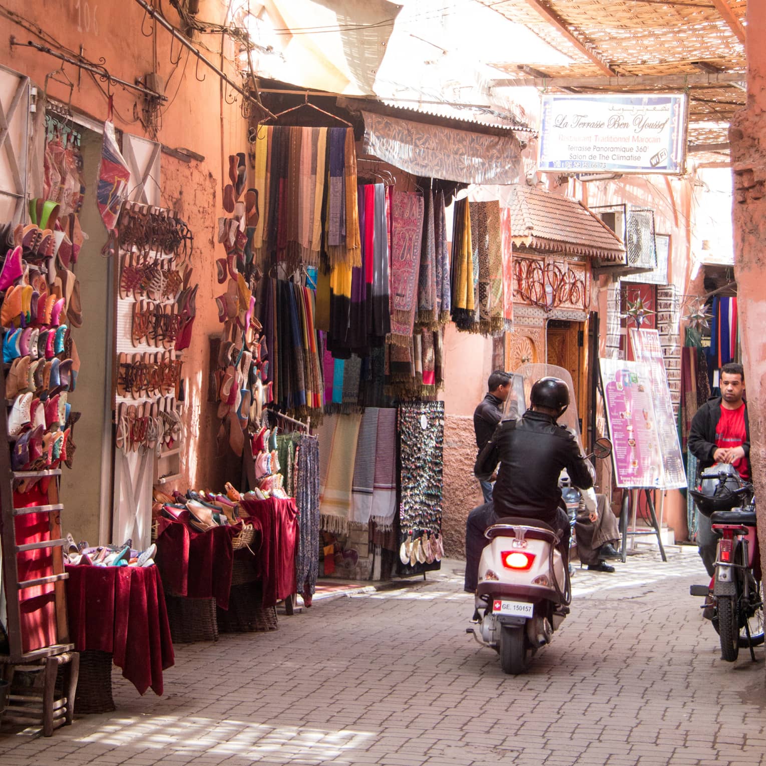 Rear view of leather-clad moped rider navigating narrow alley with colourful market items on display, wicker roof overhead.