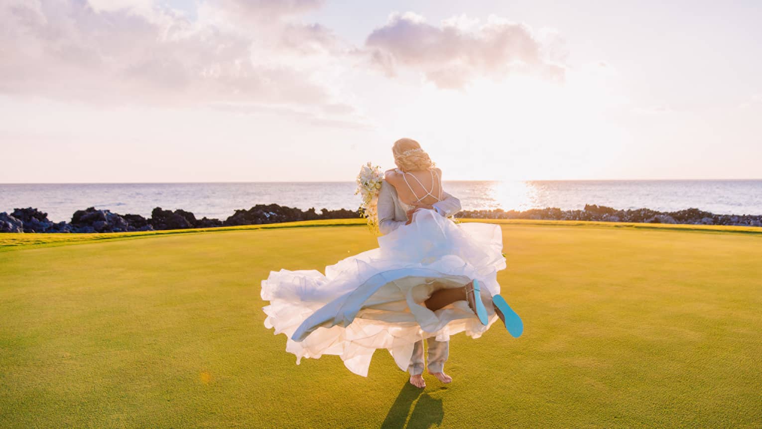 Groom lifting bride off ground on green lawn near ocean
